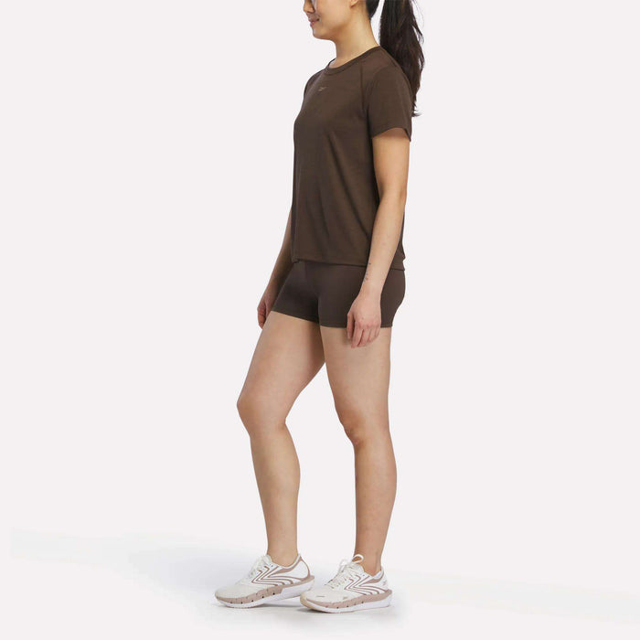 A person stands slightly sideways, wearing a Reebok Women's Training T-Shirt with matching brown shorts and white athletic shoes, set against a plain white background.