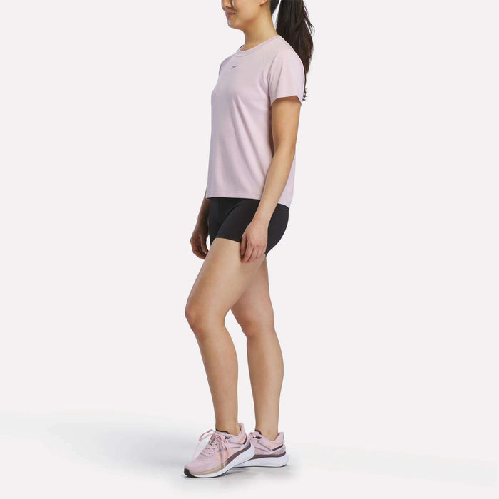 A woman stands sideways in a light pink Reebok Women's Training T-Shirt, black shorts, and pink athletic shoes. Her hair is tied back in a ponytail as she poses against a plain white background.