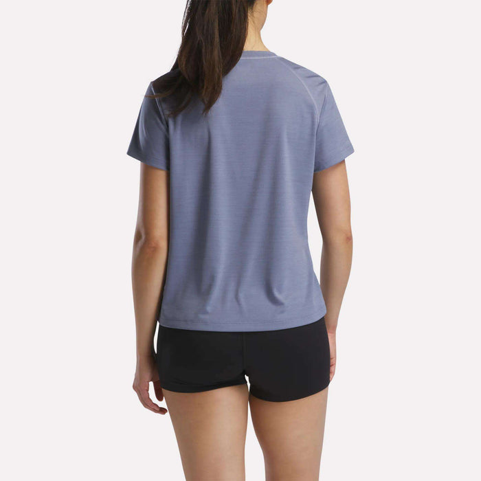 A woman with long brown hair in a ponytail stands with her back to the camera, wearing a light blue Reebok Women's Training T-Shirt and black shorts against a plain white background.