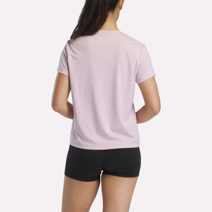 A woman with long dark hair in a ponytail is seen from the back, wearing a light pink Reebok Women's Training T-Shirt and black shorts, standing against a plain white background.