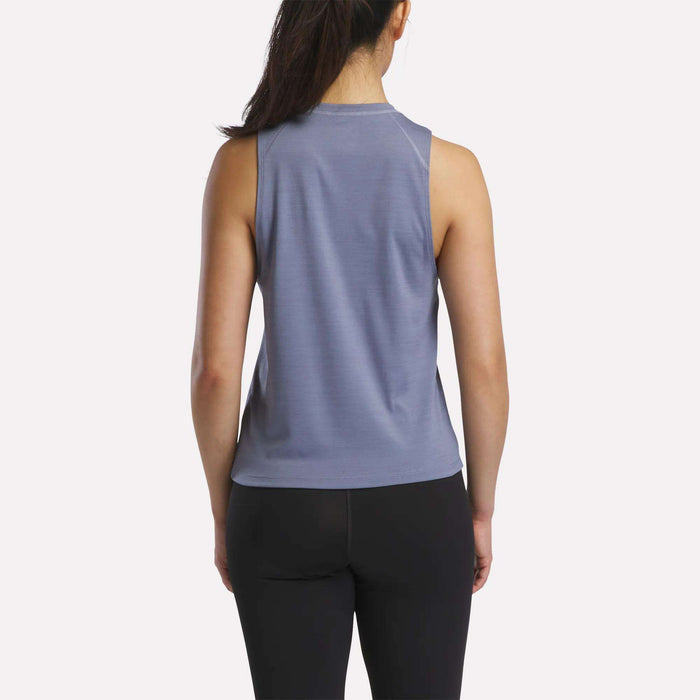 A woman with long dark hair in a ponytail is shown from behind, wearing a sleeveless blue Reebok Women's Training Tank Top and black leggings against a plain white background.
