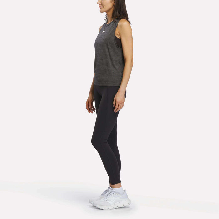 A woman stands in profile against a plain white background, wearing the Reebok Women's Training Tank Top—a sleeveless gray design with RBK-DRY technology—paired with black leggings and white athletic shoes.