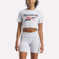A woman wears a Reebok Women's Two-Color Logo Crew Cropped Short Sleeve Tee in light gray with matching high-waisted bike shorts, standing against a plain white background.
