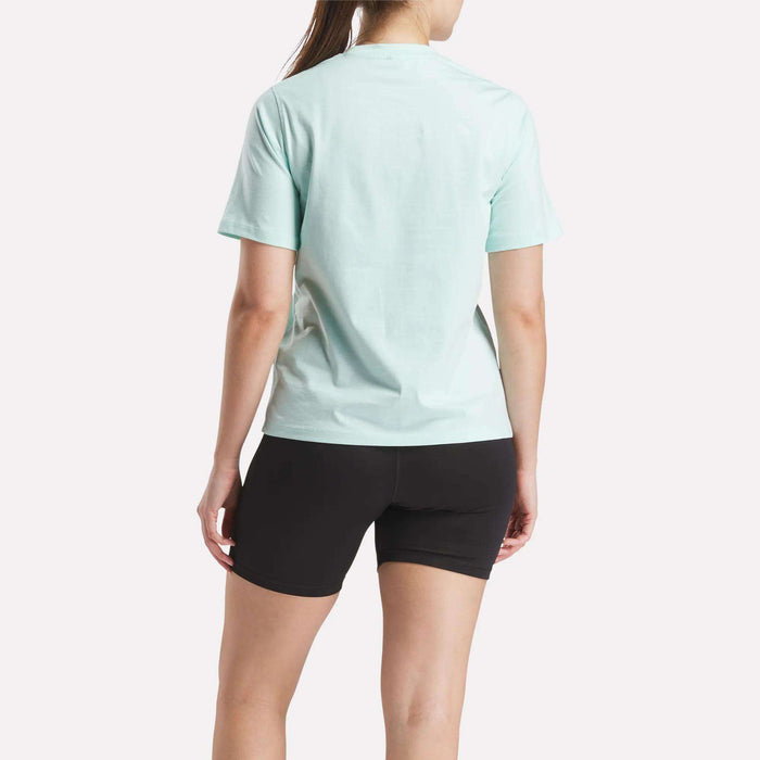 A person stands facing away, wearing a light blue Reebok Women's Two-Color Logo Crew Short Sleeve Tee and black shorts against a plain white background.