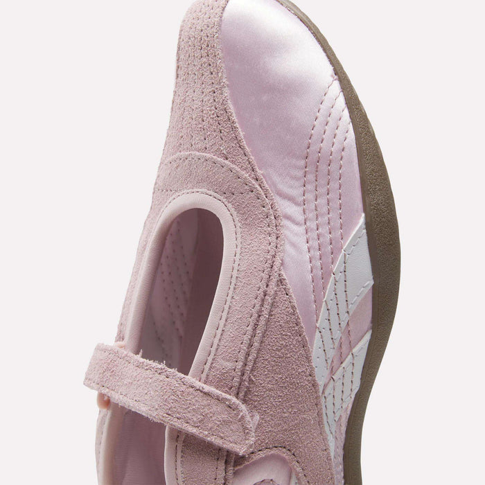 A close-up of the Reebok Women's Ultra Lo Mary Jane Shoes in pastel pink features a velcro strap, suede and satin-like textures, white side stripes, and a brown rubber sole.