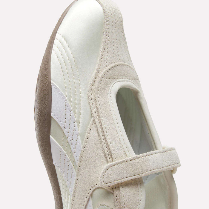A close-up of the Reebok Women's Ultra Lo Mary Jane Shoes in white and beige, featuring a hook-and-loop strap, suede and leather accents, and a textured brown sole, shown against a light background for a modern, low-profile runner look.