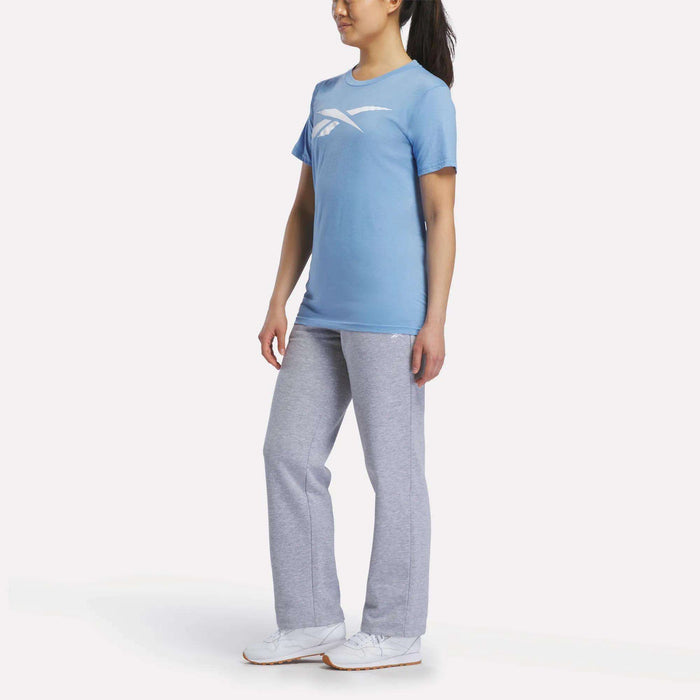 A woman stands at an angle wearing a light blue Reebok Women's Vector Graphic T-Shirt, gray sweatpants, and white sneakers against a plain white background.