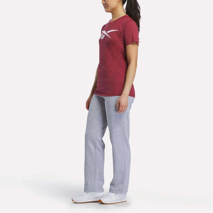A woman faces slightly to the side in a Reebok Women's Vector Graphic T-Shirt, gray sweatpants, and white sneakers against a plain white background.