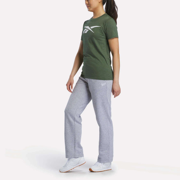 A woman wearing a green Reebok Women's Vector Graphic T-Shirt, light gray sweatpants, and white Reebok sneakers stands against a plain white background.