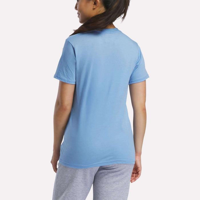 A woman with long dark hair in a ponytail stands facing away, wearing a light blue Reebok Women's Vector Graphic T-Shirt and light gray pants, set against a plain white background.