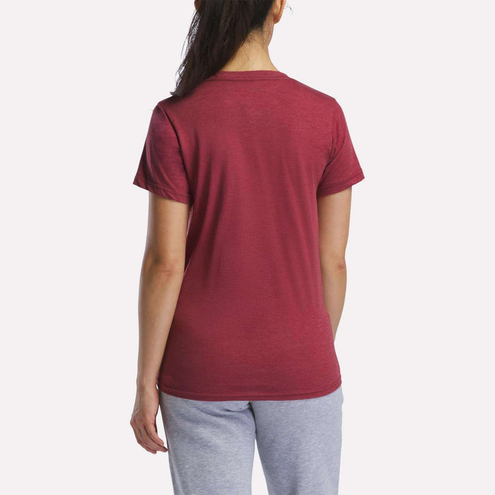 A woman with long dark hair in a ponytail is seen from the back, wearing a Reebok Women's Vector Graphic T-Shirt in maroon with light gray pants, standing against a plain white background.