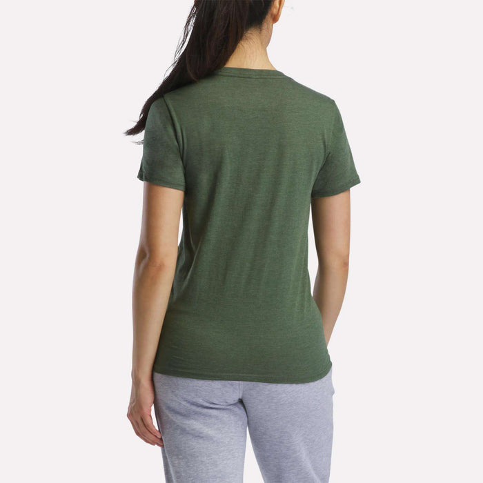 A woman with long dark hair in a ponytail is seen from behind, wearing a green Reebok Women's Vector Graphic T-Shirt and light gray sweatpants, standing against a plain white background.
