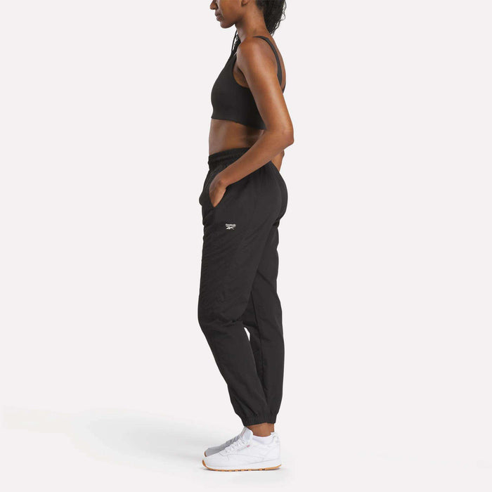 A woman wears Reebok Women's Vector Track Pants in black, paired with a sports bra and white sneakers, standing in profile against a plain white background with her hands in her pockets.