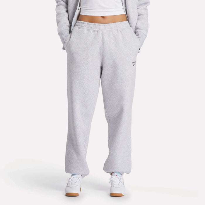 A person wearing Reebok Women's Wardrobe Essentials Sweat Pants with hands in pockets, paired with a white cropped top, white sneakers, and a matching gray jacket. The background is plain white.