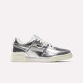 A pair of Reebok Women’s Workout Plus Shoes in Silver Metallic/Chalk/Vintage Chalk, crafted from premium garment leather with matching white laces and chalk-toned sole, shown from a side angle against a plain white background for a classic sneaker aesthetic.