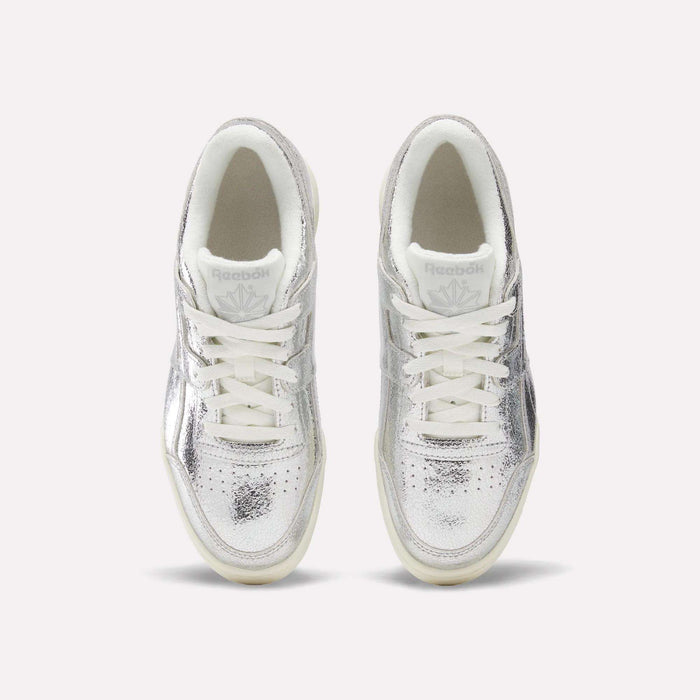 Here’s a rewritten version using the provided product data: A top-down view of Reebok Women’s Workout Plus Shoes crafted in premium garment leather, showcasing their shiny silver finish with white laces and soles, elegantly positioned side by side against a plain light background.