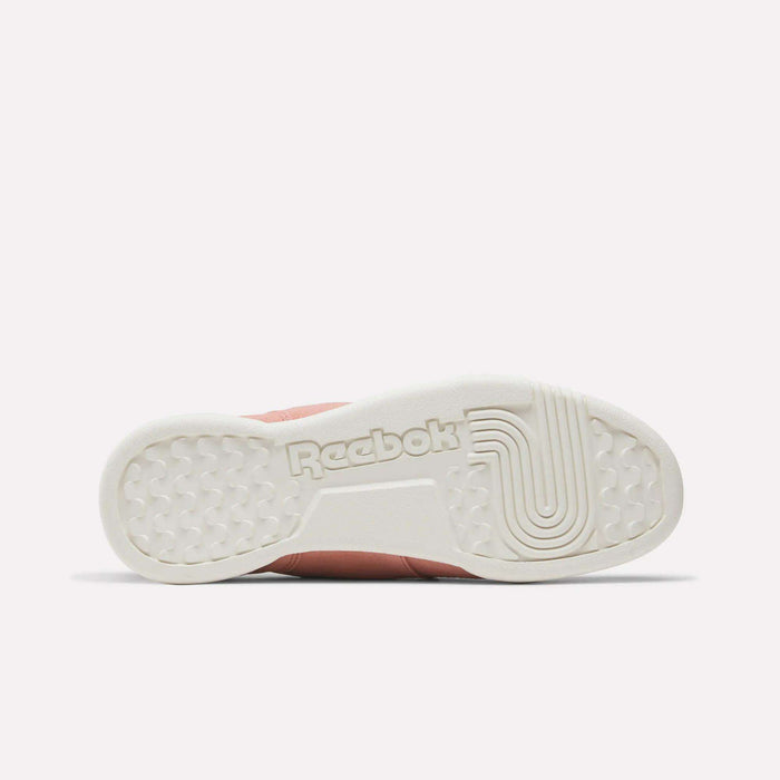 The image displays the bottom view of Reebok Women's Workout Plus Shoes, highlighting the white sole with the Reebok logo, unique tread for grip and support, and a muted pink upper that channels an 80s fitness vibe.