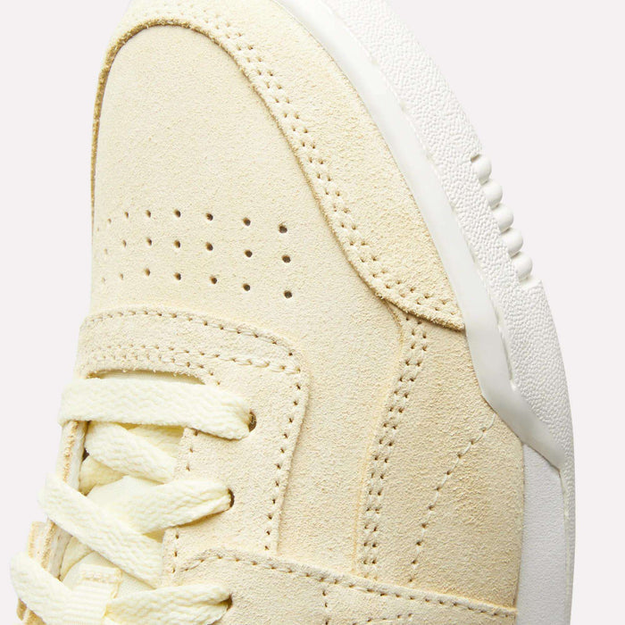 Close-up of the upper front of the Reebok Women's Workout Plus Shoes, featuring beige suede, double perforations, cream laces, and a white textured rubber sole—a classic '80s-inspired fitness style.
