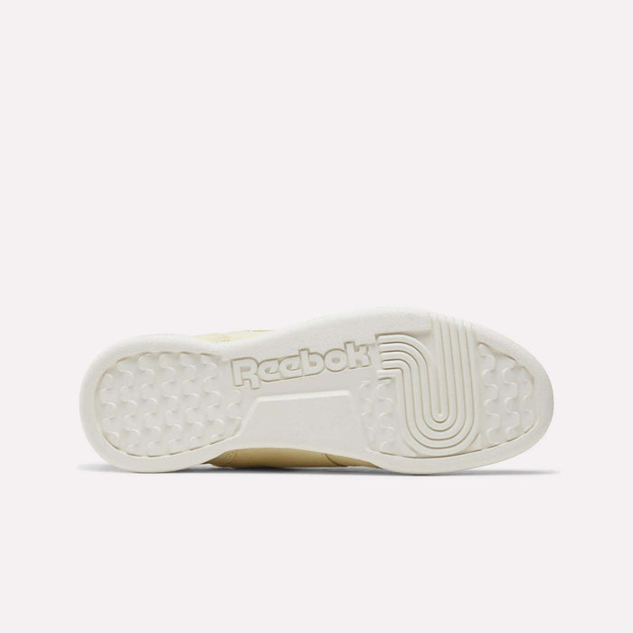 The image displays the outsole of the Reebok Women's Workout Plus Shoes in off-white, highlighted by textured grip patterns and a prominent Reebok logo at the center, reflecting classic '80s fitness style.