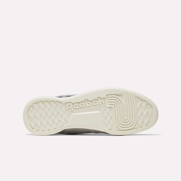 The image features the sole of the Reebok Women’s Workout Plus Shoes, laid out horizontally. The outsole appears off-white with intricate textured patterns, and “Reebok” is prominently embossed at the center, showcasing the classic sneaker’s detail-oriented design and quality craftsmanship.