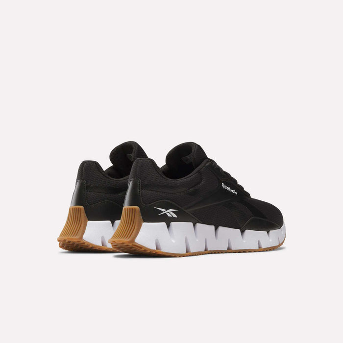 A pair of Reebok Women's Zig Dynamica STR shoes in black, featuring unique white zigzag midsoles and brown rubber outsoles, are displayed on a light background showing the side and back angles.