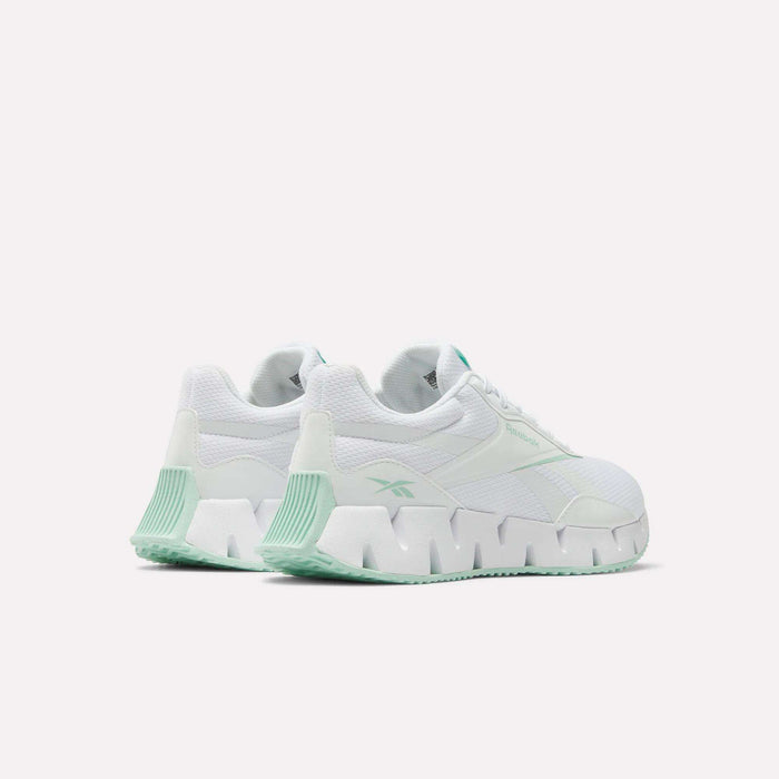 The Women's Zig Dynamica STR Shoes by Reebok feature chunky geometric soles, breathable mesh panels, and light green accents on white uppers. Innovative running shoes are shown against a plain white background.