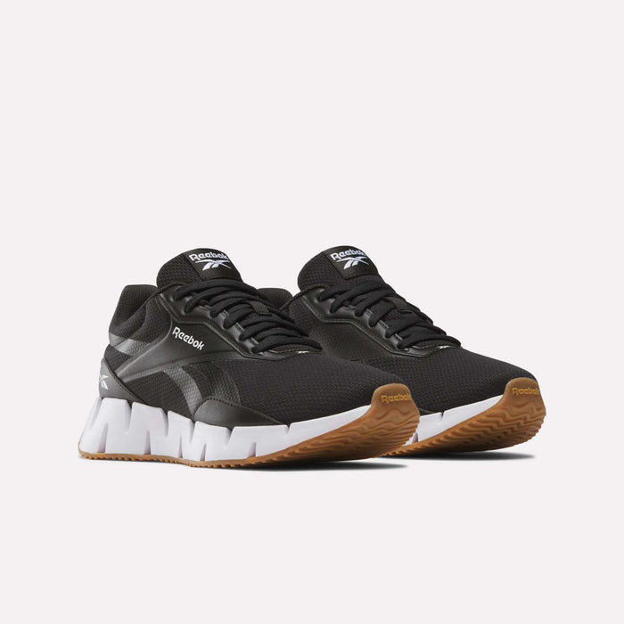 A pair of Reebok Women's Zig Dynamica STR Shoes in black with white zigzag soles and brown outsoles, designed for superior grip, displayed on a plain light background.