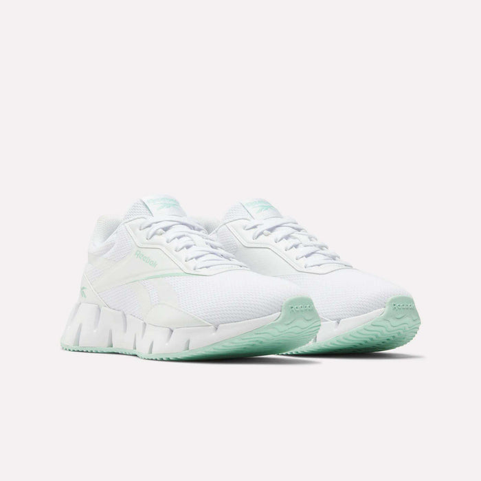A pair of Reebok Women's Zig Dynamica STR Shoes in white, featuring breathable mesh and light mint green accents on the soles and branding, displayed on a white background.
