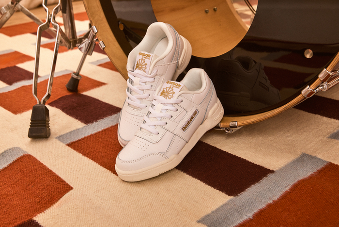 A pair of white Reebok sneakers with gold accents, resting on a patterned rug next to a drum set.