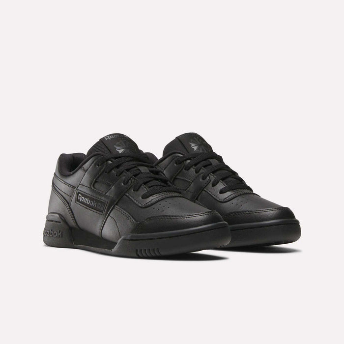 A pair of black Reebok Workout Plus Shoes for big kids, featuring matching black soles and laces with the Reebok logo on the side and tongue, displayed against a plain white background.