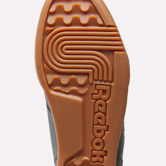 Close-up view of the Reebok Workout Plus Shoes for Big Kids, highlighting a gum rubber outsole with wavy tread patterns and the Reebok logo embossed in large letters.