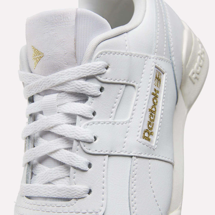 A close-up of Reebok Workout Plus Shoes for big kids, showcasing white textured leather, gold Reebok logos on the tongue and side, and crisp white laces.