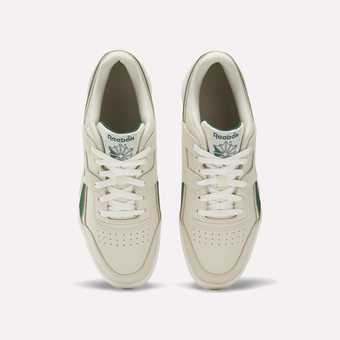 A top-view image of a pair of women’s Reebok Workout Plus Shoes in off-white leather with green details, displayed on a plain light background. The sneakers feature laces, perforated toe boxes, and tongue tags showcasing the Reebok logo and green leaf.