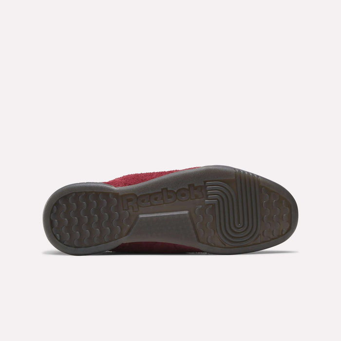 The image shows the bottom view of Reebok Workout Plus Shoes, featuring a dark rubber sole with grip patterns and the Reebok logo. The upper, inspired by ’80s fitness style, is partly burgundy.