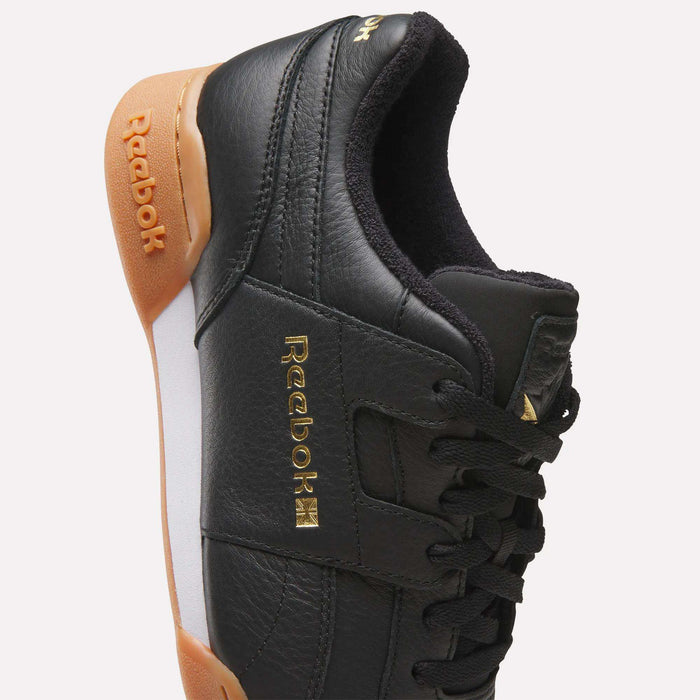 A side-profile view of the Reebok Workout Plus Shoes highlights their classic ’80s fitness style, featuring black leather uppers, gold Reebok logos, a contrasting white midsole panel, gum outsole, and black laces—all set against a plain background.