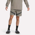 A person in a gray long-sleeve shirt, Reebok Woven Basketball Shorts, black socks, and black slip-on shoes stands against a plain background with arms relaxed at their sides.