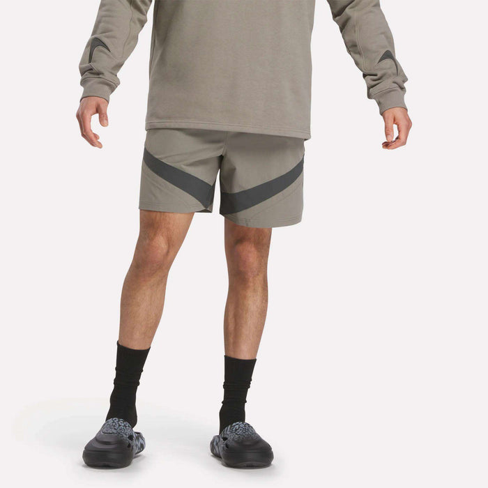 A person in a gray long-sleeve shirt, Reebok Woven Basketball Shorts, black socks, and black slip-on shoes stands against a plain background with arms relaxed at their sides.