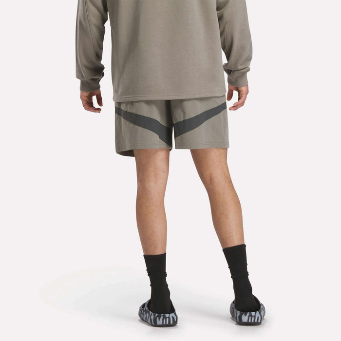 The lower half of a person is shown wearing a taupe long-sleeve shirt, Reebok Woven Basketball Shorts in gray with dark chevron stripes, black socks, and gray patterned slip-on shoes.