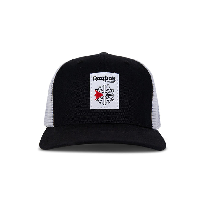 The Reebok Woven Patch Vector Trucker Cap features a black and white design, curved brim, mesh back, adjustable snapback, and a front woven patch with the Reebok logo plus a bold red accent.