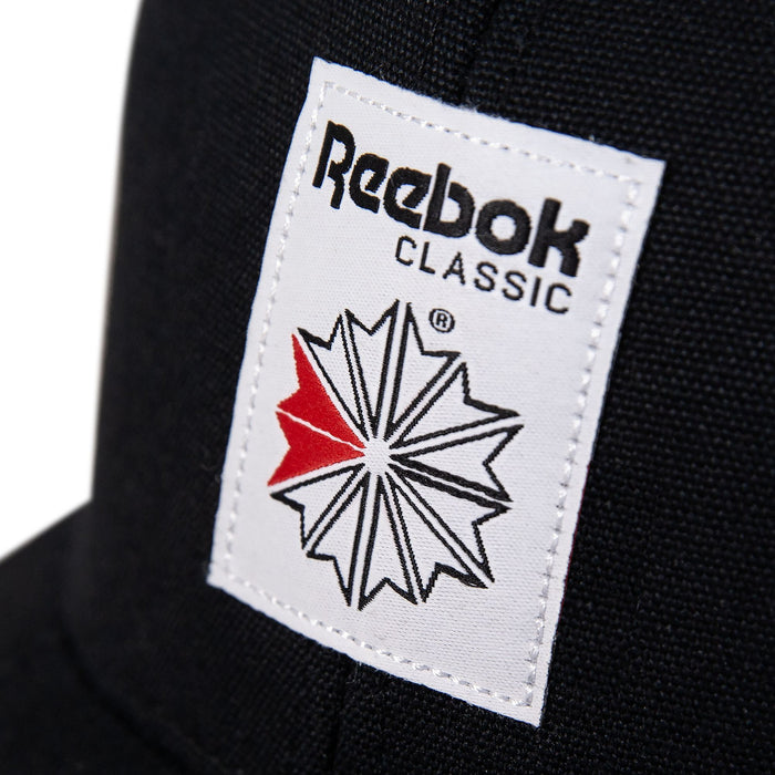 Close-up of the Reebok Woven Patch Vector Trucker Cap in black, featuring a white woven patch with the Reebok Classic logo, a geometric star design with red and black accents, and an adjustable snapback for comfort.