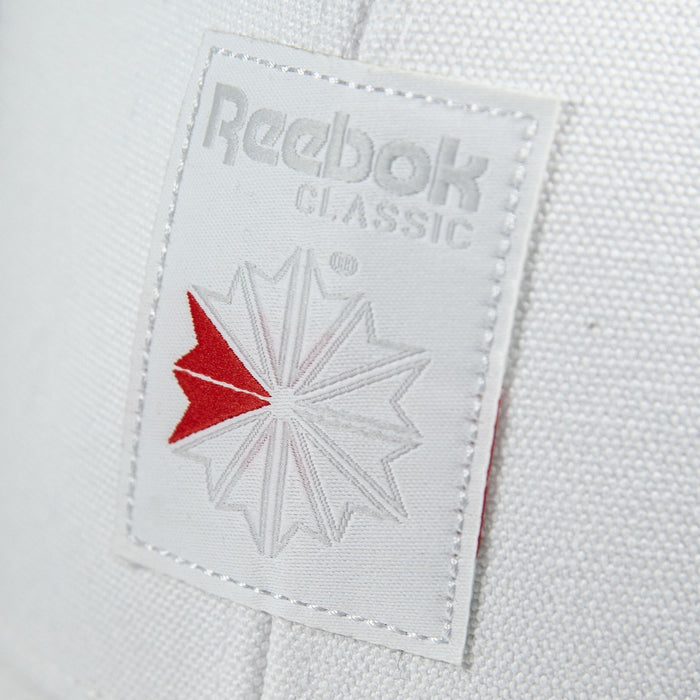 Close-up of the Reebok Woven Patch Vector Trucker Cap, showing a white Reebok patch with the brand name and starburst logo. The cap features mesh back panels and an adjustable snapback closure.