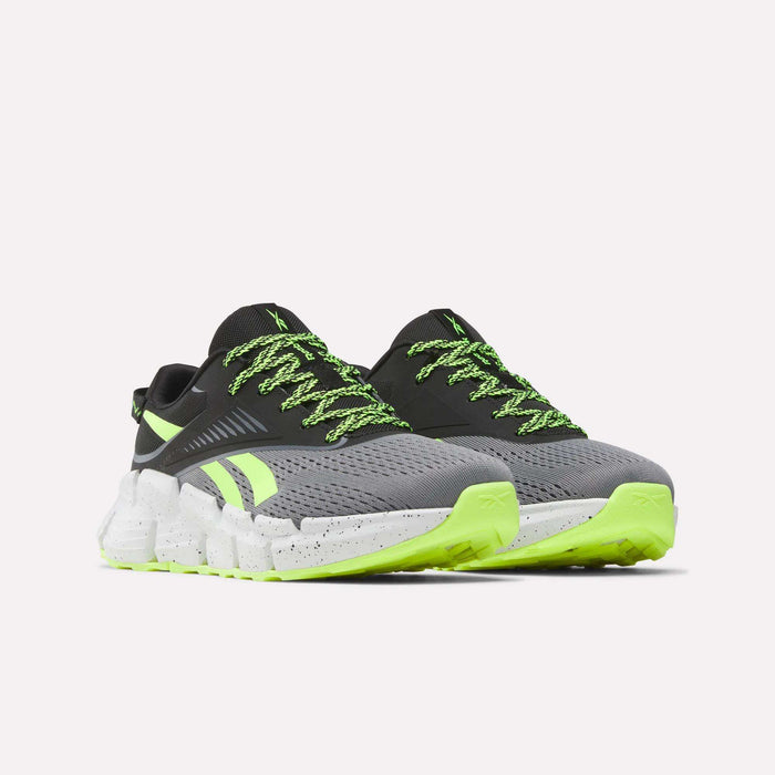 A pair of Reebok Zig Dynamica 6 Running Shoes for big kids, featuring grey mesh uppers, black accents, neon green laces, and white chunky soles with black speckles—shown on a plain white background.
