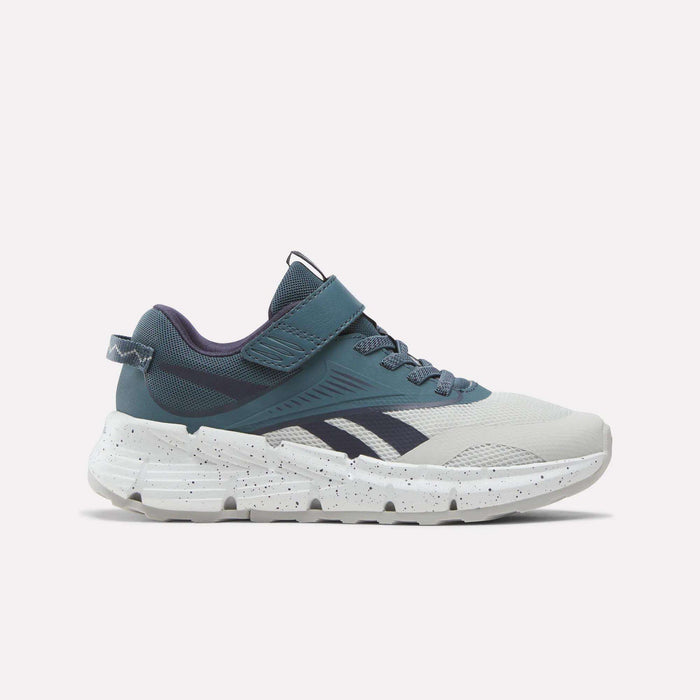 A pair of Reebok Zig Dynamica 6 Running Shoes for little kids is shown in side profile on a plain white background, featuring a dark indigo, midnight tide, and new grey 1 colorway. These athletic sneakers showcase Reebok’s Zig technology with a thick speckled sole, navy accents, mesh panels, and a Velcro strap.